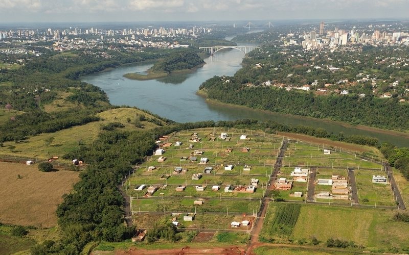 Vista aérea do loteamento: quadras demarcadas em primeiro plano e, ao fundo, a Ponte da Amizade sobre o rio Paraná com o skyline de Foz do Iguaçu.