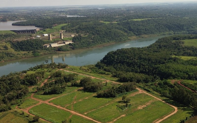 Vista aérea do loteamento com as quadras em campo verde e, ao fundo, a barragem de Itaipu e o rio Paraná.